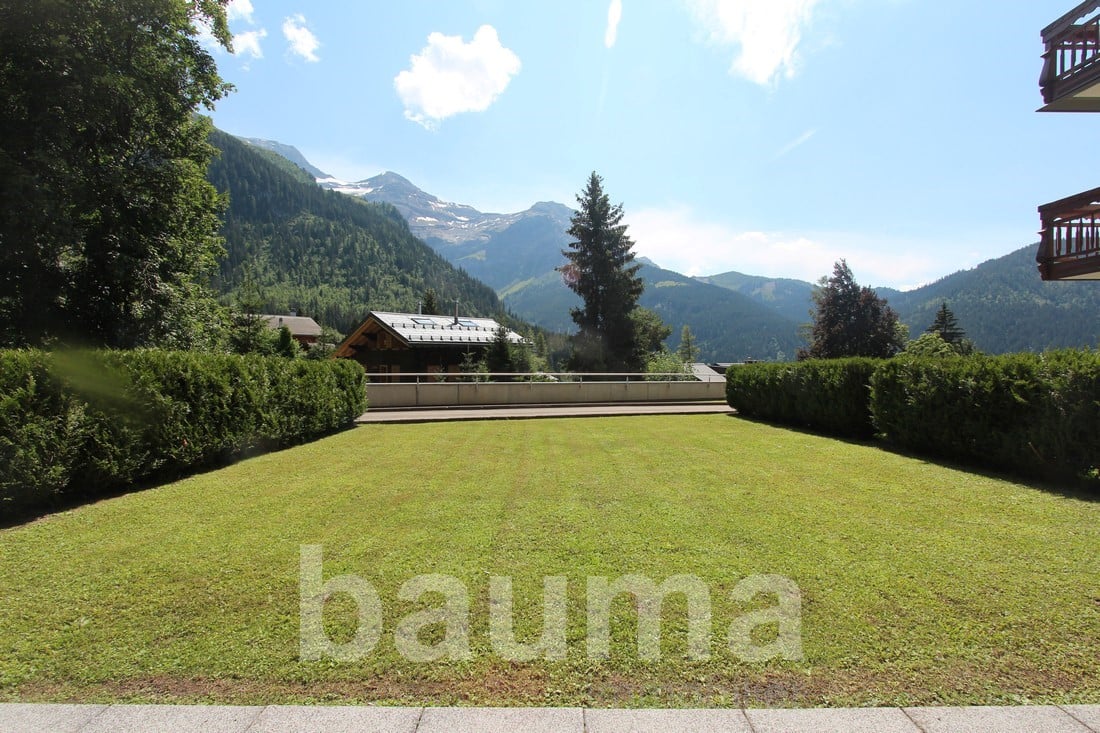 The image shows a scenic mountain landscape with a large grassy lawn in the foreground. There are several wooden chalets or cabins visible, with balconies or terraces. The mountains in the background are covered in lush green forests. The sky is blue with