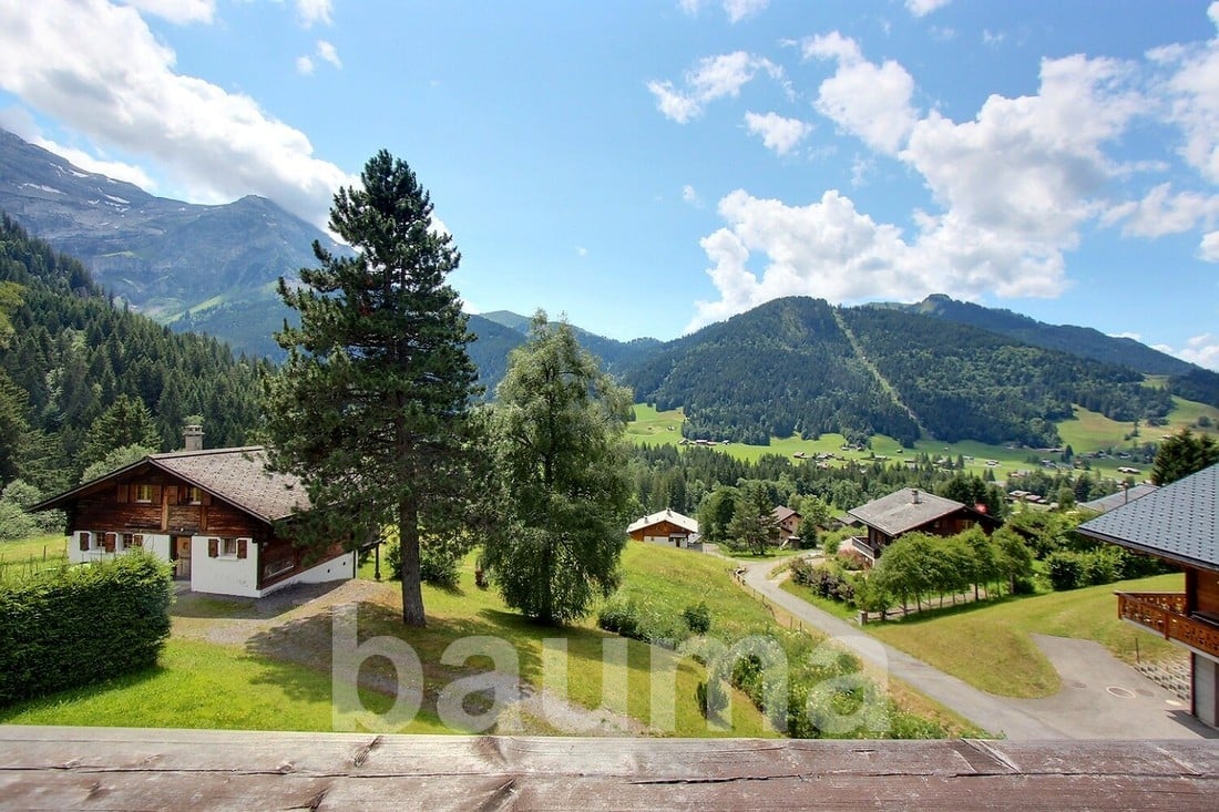 The image shows a scenic mountain landscape with lush green forests, rolling hills, and traditional alpine-style houses. The sky is blue with fluffy white clouds, creating a picturesque and serene atmosphere. The landscape appears to be in a rural, mounta