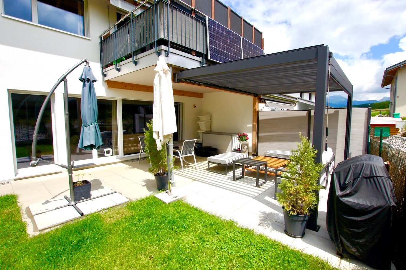 2-story modern house with balcony, covered patio area with outdoor furniture, solar panels on the roof, lawn, and various potted plants