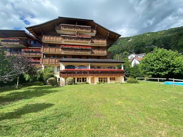 multi-story house with balconies, large grassy area in front, surrounded by trees and bushes, mountains in the background