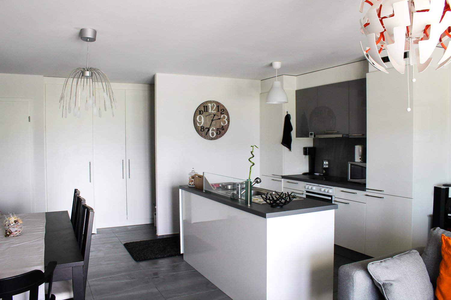 Modern kitchen with a large island, white cabinets, black countertops, modern fixtures, a wall clock, and white tiles.
