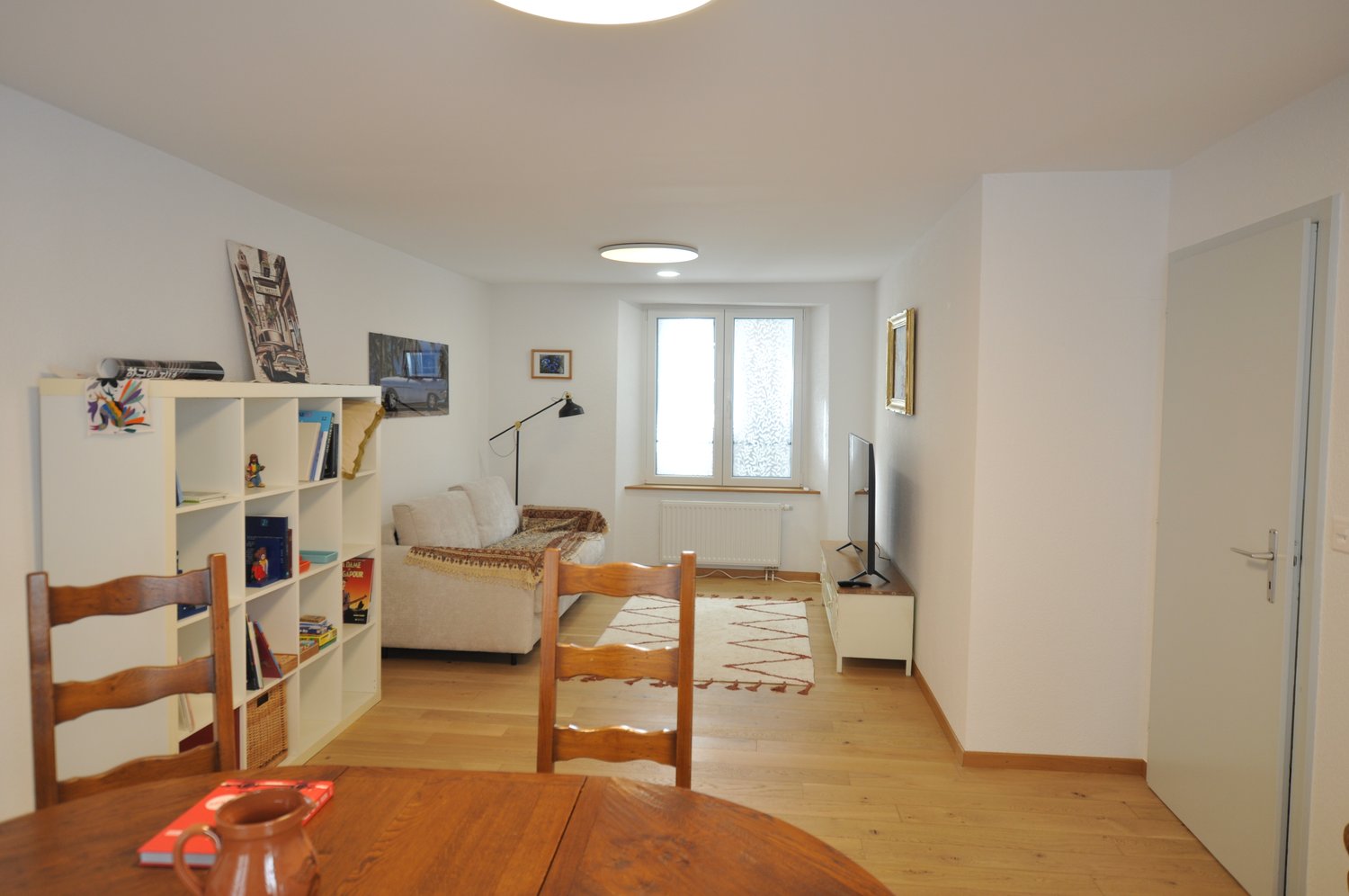 Spacious living room with hardwood floors, white walls, large windows, bookshelf, sofa, coffee table, and artwork on the walls.