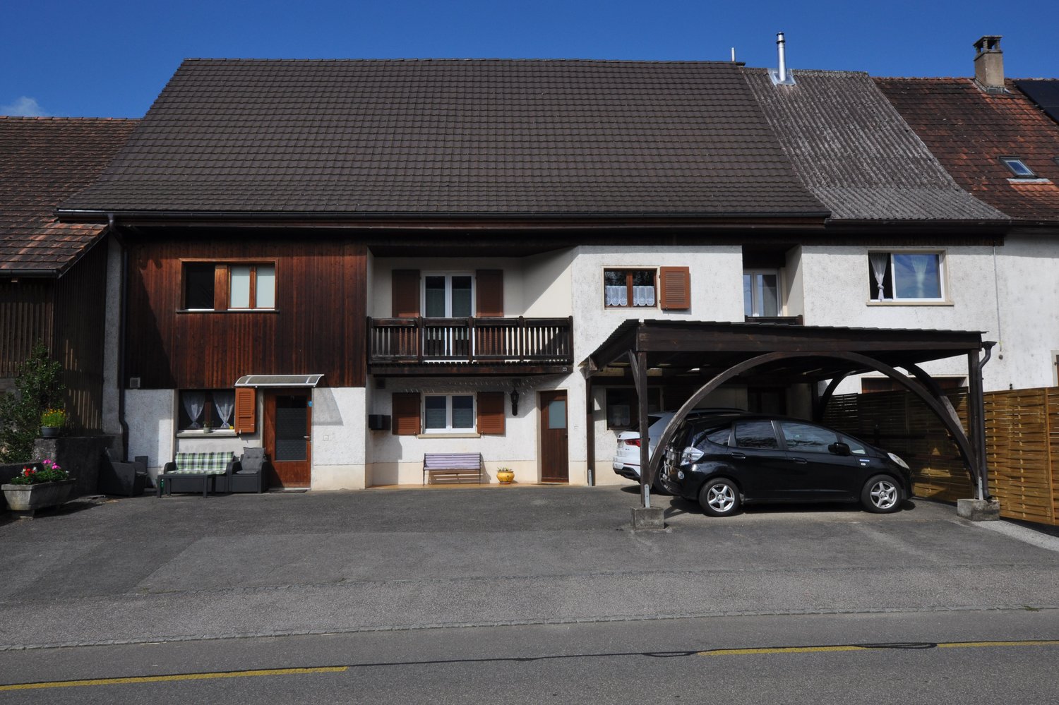 2 story house, brown roof, balcony, wooden fence, 2 parked cars, 2 cars sheltered by carport, garden bench, sofa, chairs