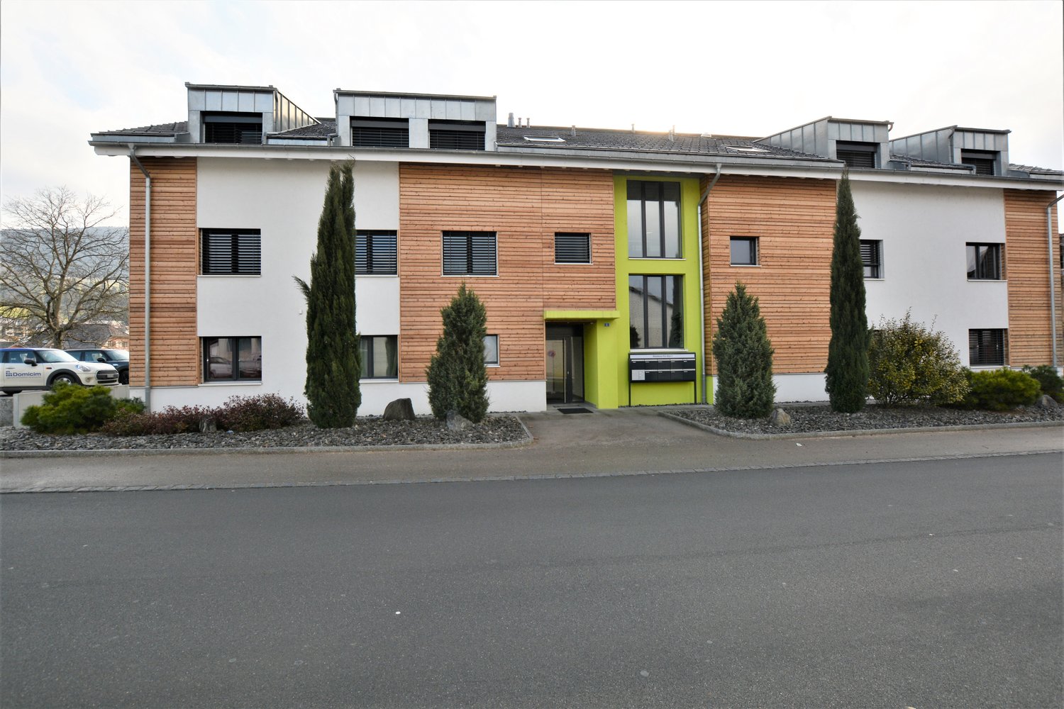 Modern apartment building with wooden panels, white walls, and green accents, featuring multiple windows and a central entrance. The building is flanked by trees and shrubs, with a paved driveway in front and several cars parked nearby.