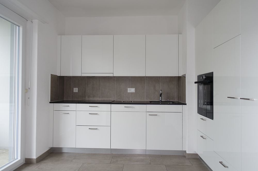 Modern, minimalist kitchen with white cabinets, dark countertops, and built-in appliances including a dishwasher. The kitchen has a clean, sleek design with ample storage space.