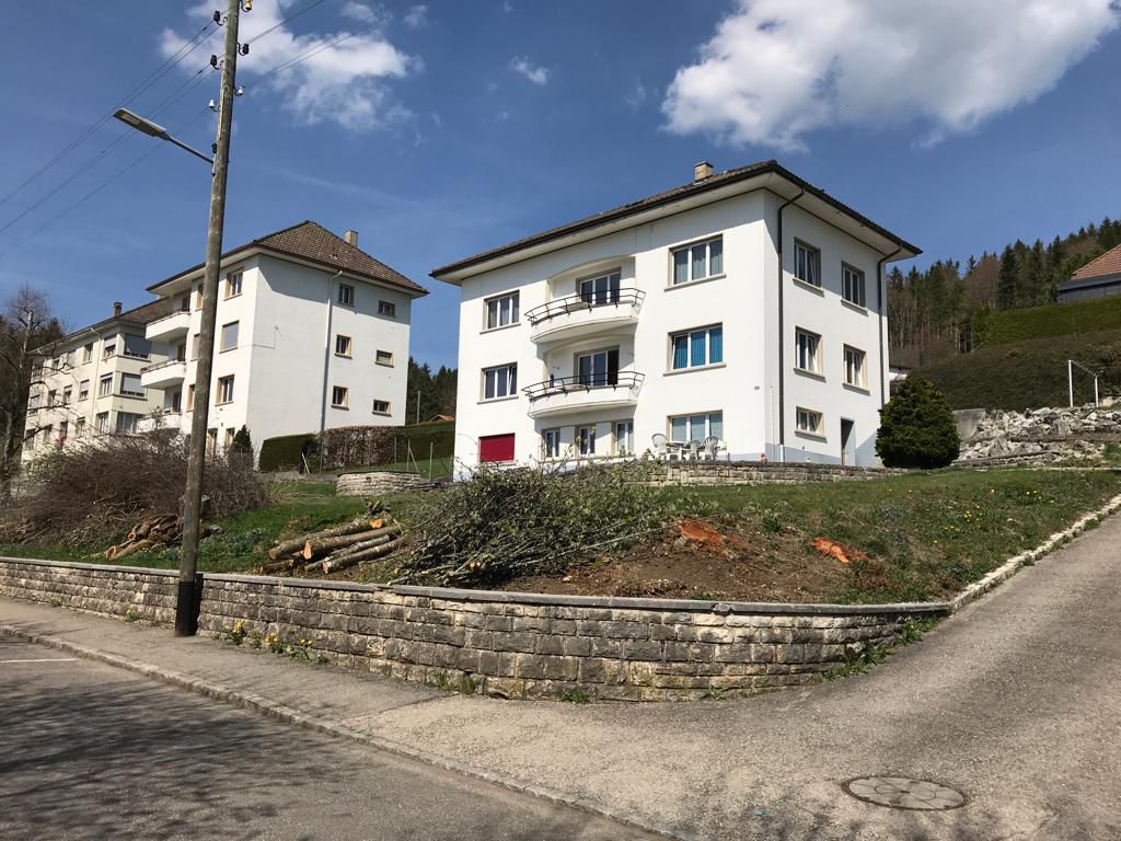 2-story white building, brown roof, balconies, red door, brick wall foundation, green grass, trees in background