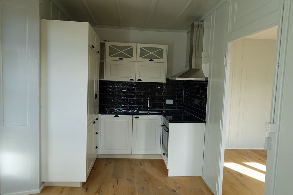 Minimalist kitchen with white cabinets, black tiled backsplash, wooden floor, integrated dishwasher, and under-cabinet lighting.