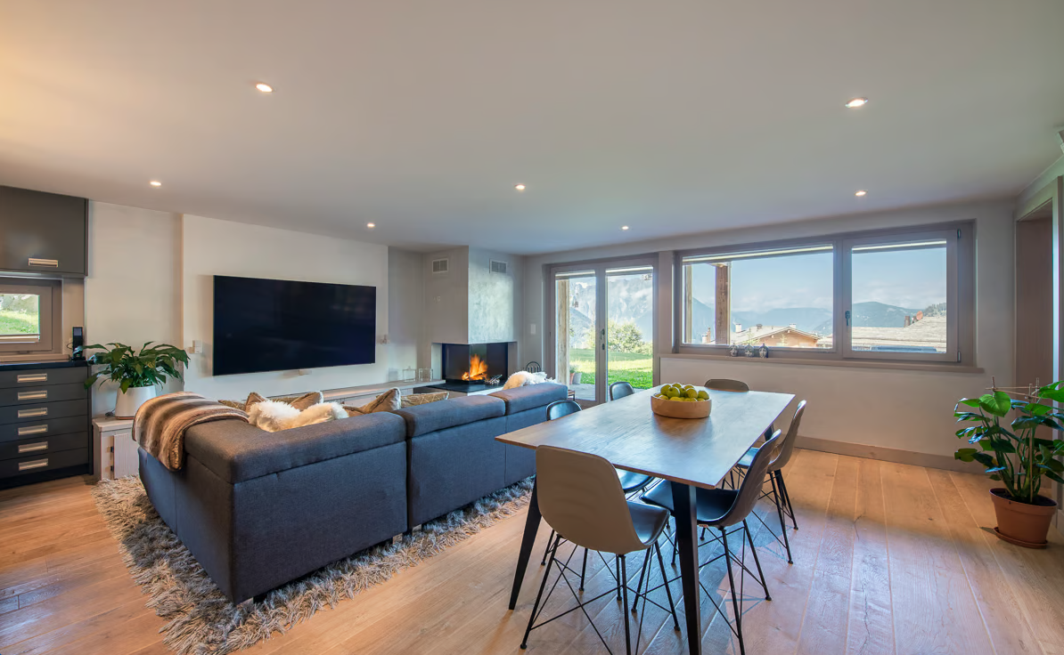 Open plan living room with L-shaped couch, fireplace, flat-screen TV, dining table, and large windows offering a mountain view.