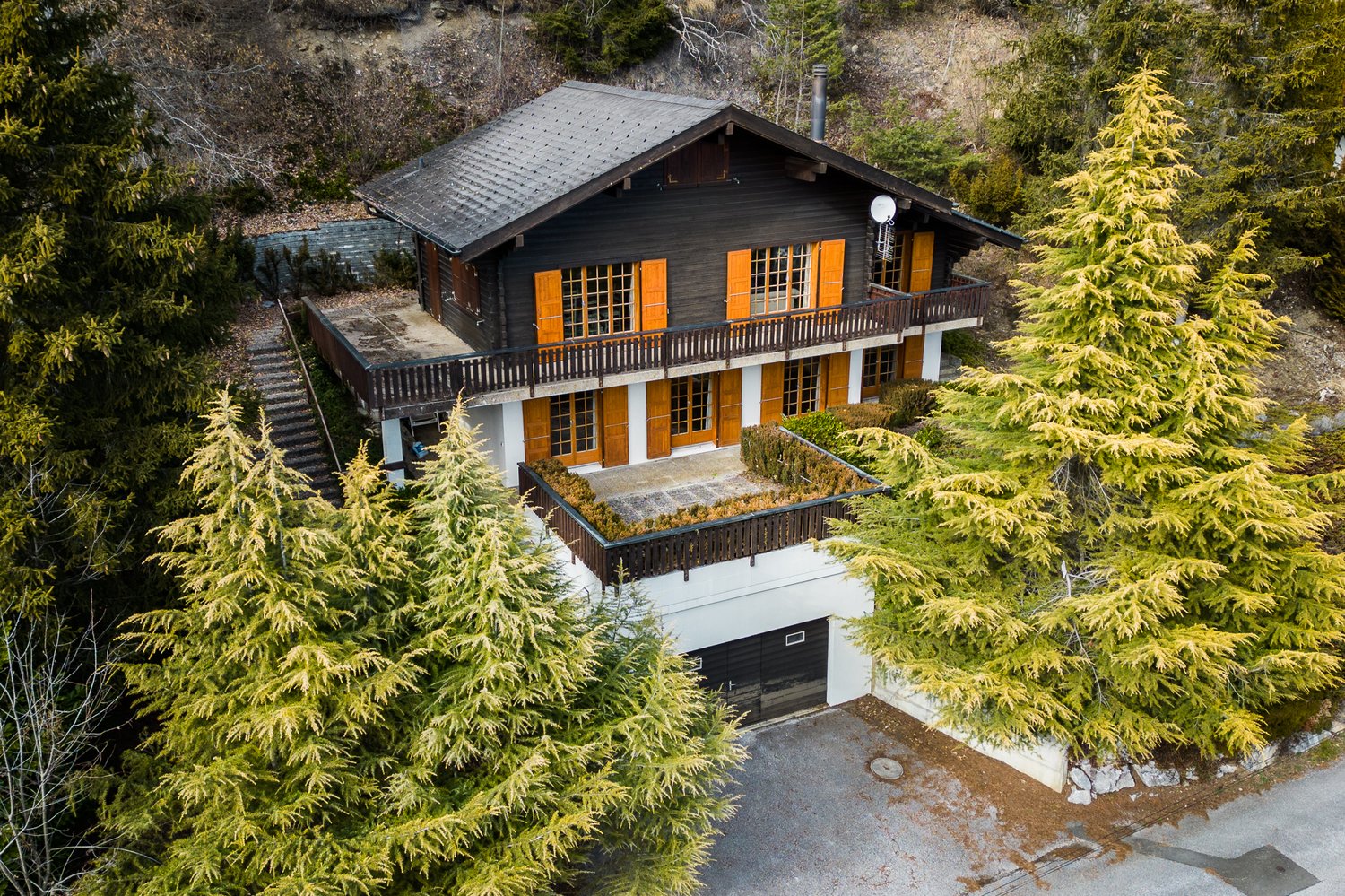 2-story wooden chalet-style house with a slanted dark roof, large windows, and a balcony/terrace. The house is surrounded by lush green pine trees and is situated in a forested, mountainous area.