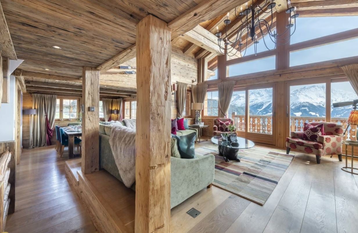 Spacious open-plan living area with wooden beamed ceiling, large windows offering mountain views, plush sofas and armchairs, a fireplace, and a chandelier lighting fixture.