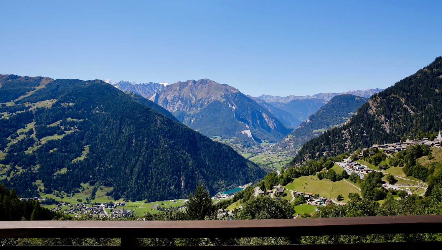 Mountain valley with lush greenery, a winding road, and distant houses, offering panoramic views of surrounding mountains.