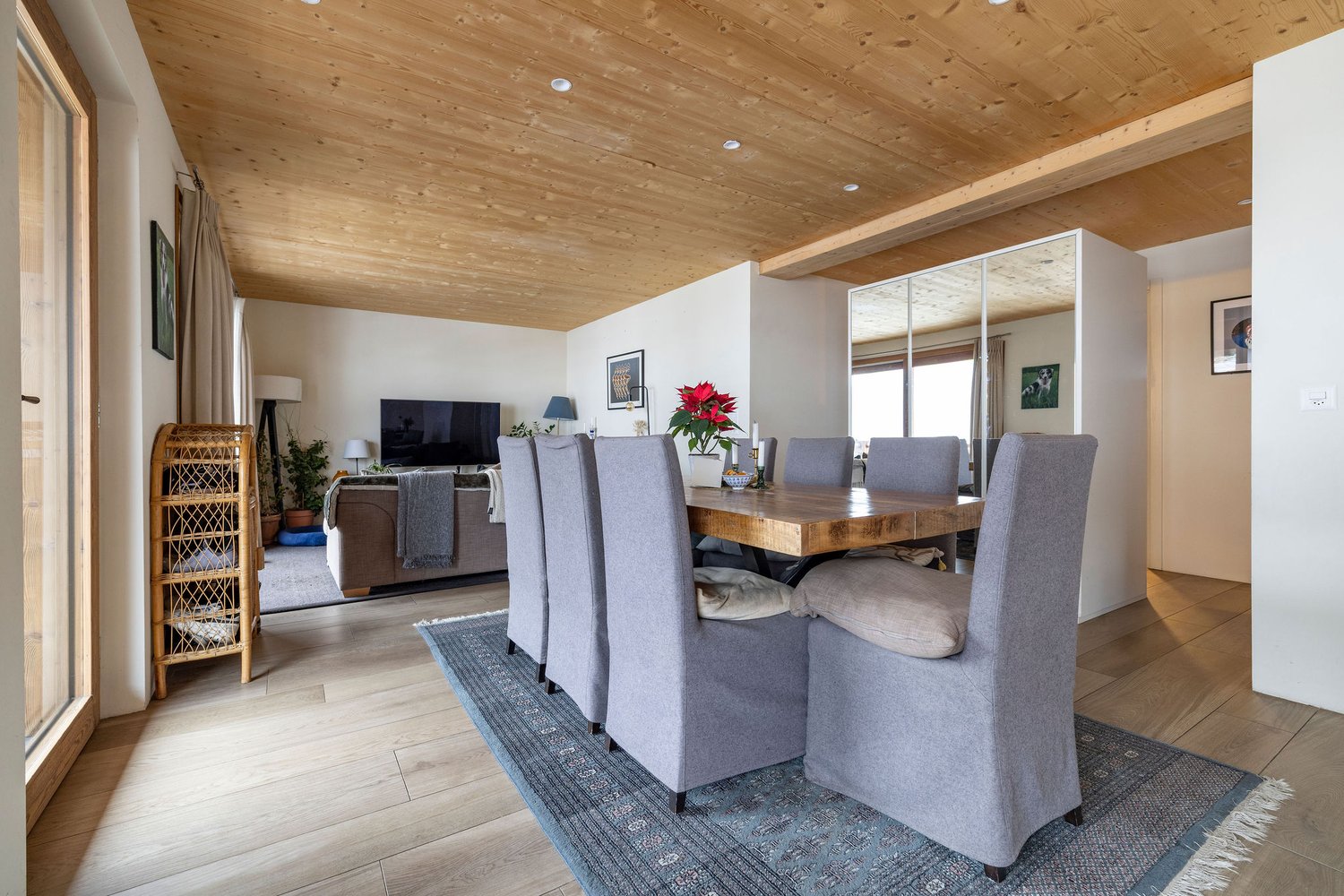 Dining room with wooden table, gray chairs, wooden ceiling, carpet, living room with couch, TV, plants, and a mirror.