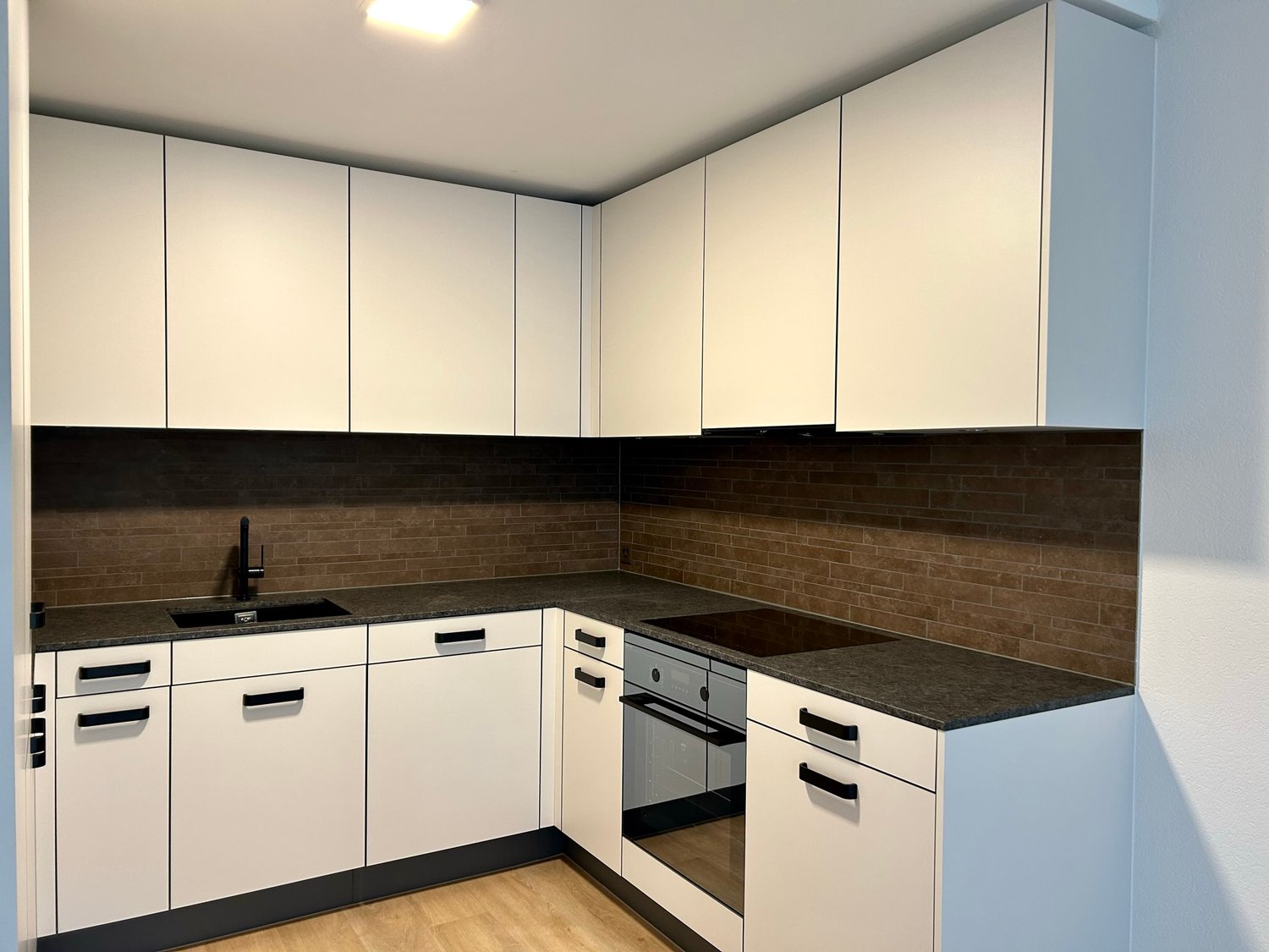 Modern kitchen with white cabinets, black countertops, and a brick backsplash. The kitchen includes a dishwasher and built-in oven.