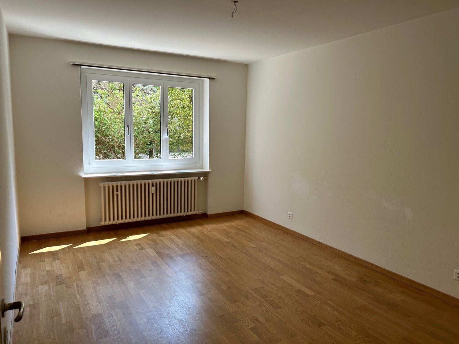 Empty room with wooden floor, white walls, large window, white radiator, power outlet