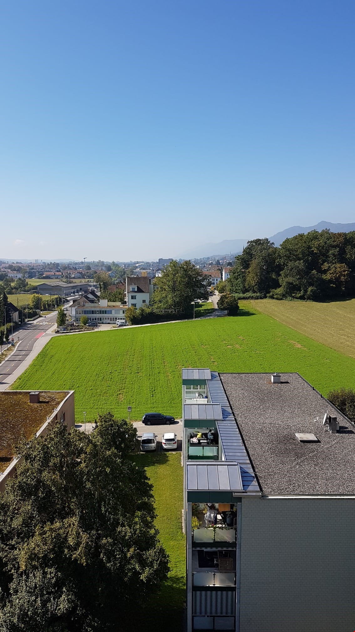 The image shows a scenic view of a residential area with a mix of buildings, green spaces, and mountains in the background. The foreground features a large grassy field, while the buildings in the middle ground have a modern, glass-and-steel design. The s