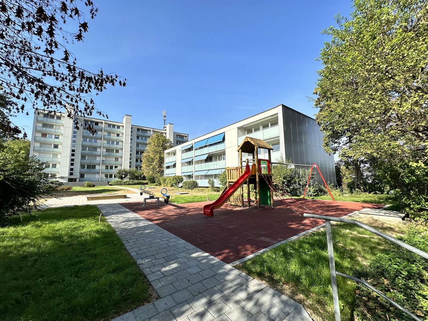 The image shows a multi-story apartment complex with balconies and a playground area in the foreground. The buildings have a modern, minimalist design with large windows. The surrounding area is well-landscaped with trees and greenery.