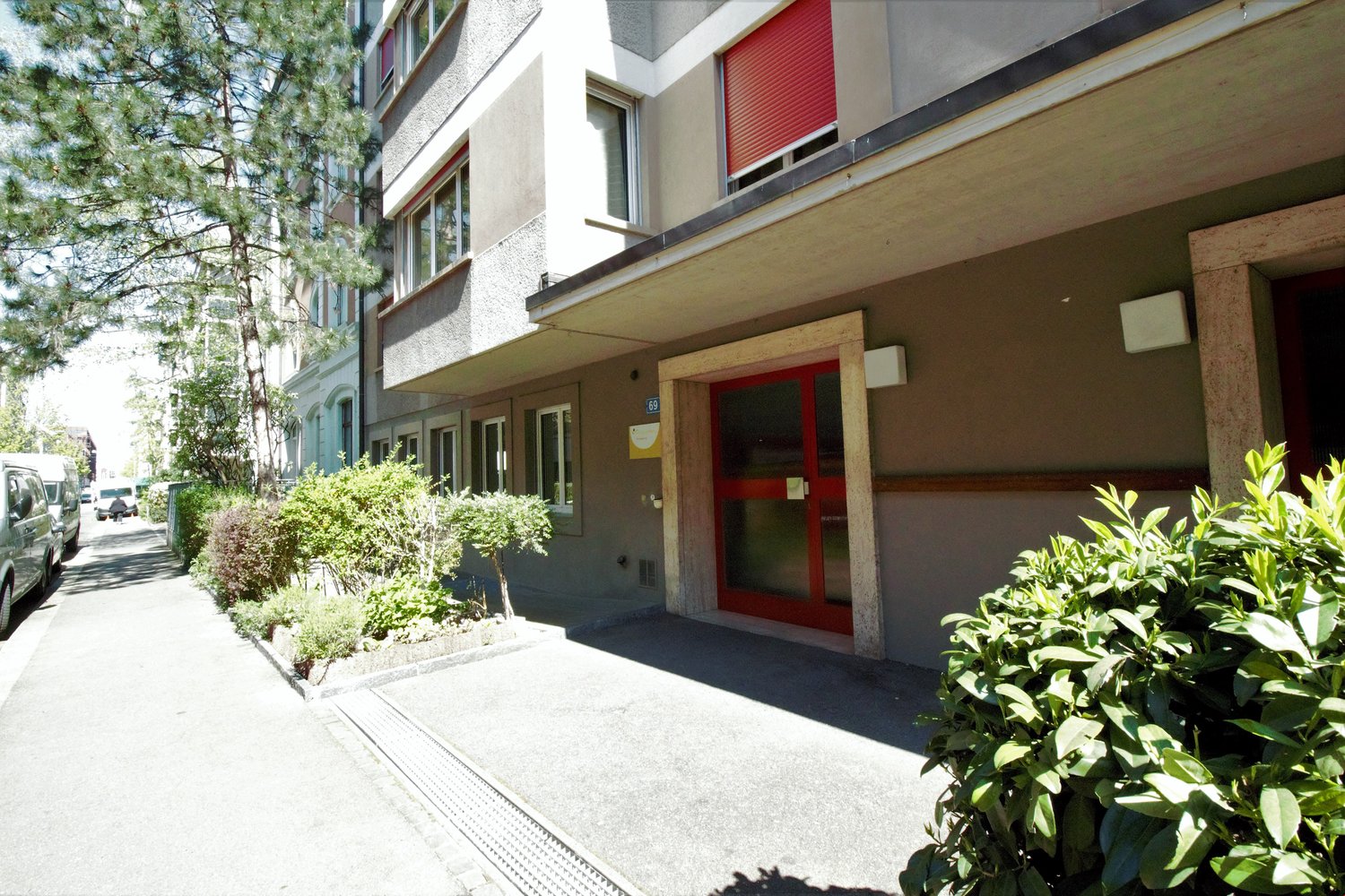 multi-story apartment building, concrete, entrance with red door, gray exterior, shrubs and trees surrounding