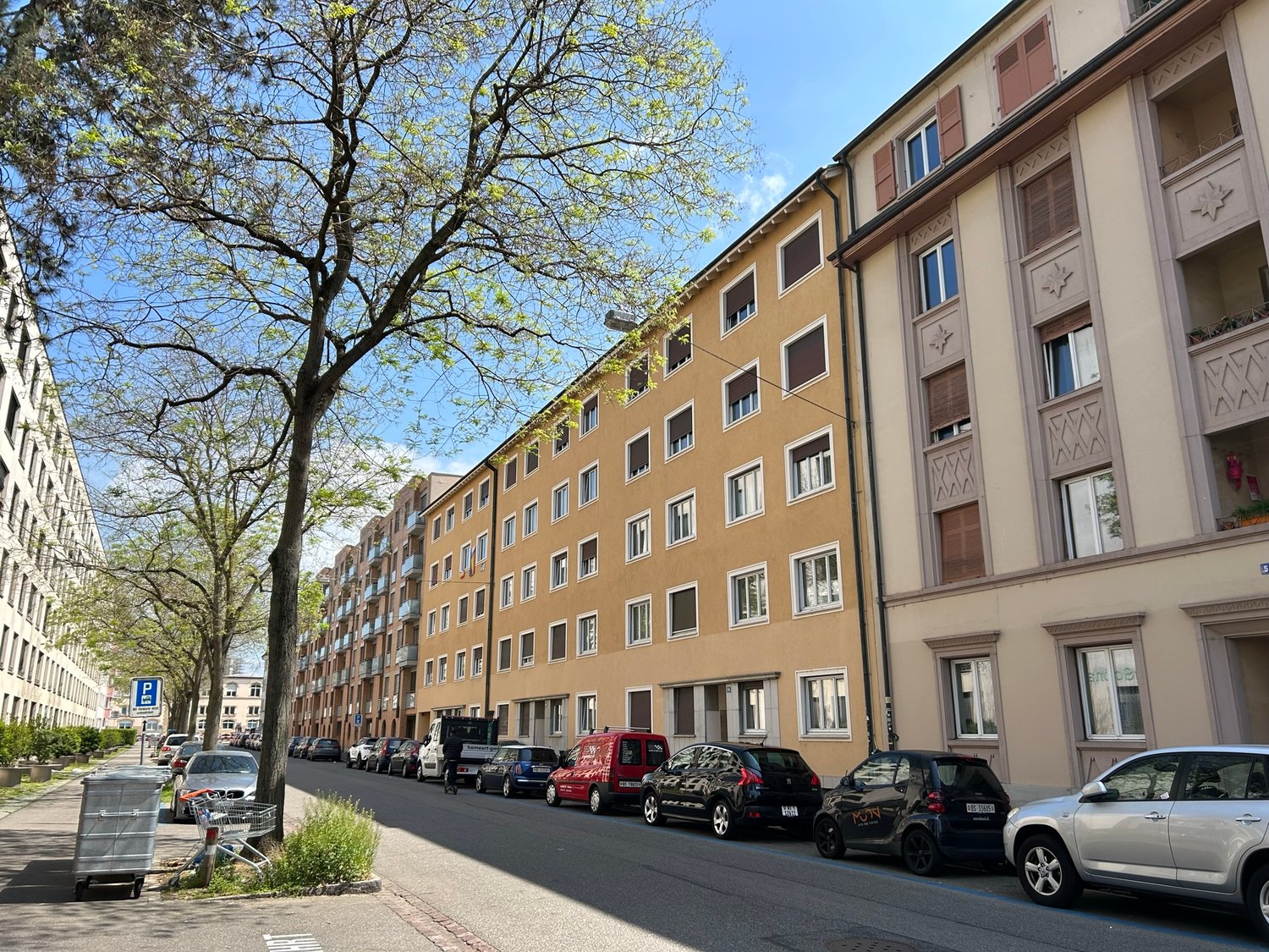 The image shows a multi-story residential building with a row of parked cars on the street in front of it. The building has a tan-colored facade with decorative architectural elements. There are trees lining the street, providing some greenery and shade.