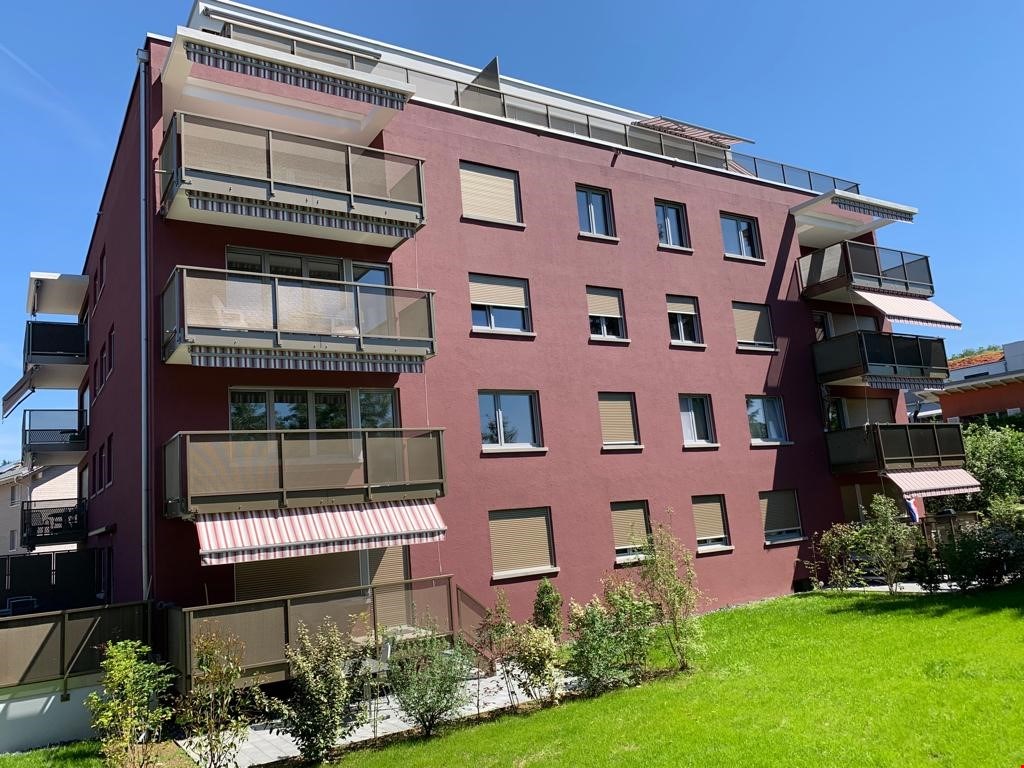 Apartment building with 4 floors, brick exterior, multiple balconies with awnings, modern design, well-maintained garden in front, red walls, glass windows.