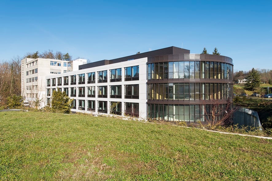 A modern, multi-story office building with a curved glass facade and a large grassy area in front. The building has multiple floors and appears to be surrounded by a parking lot or other paved area for parking.