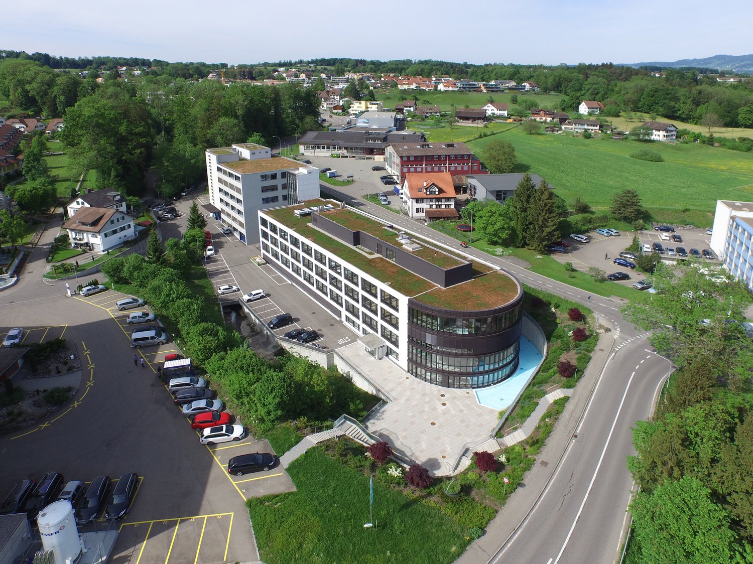 A large multi-story commercial or office building with a curved glass facade, surrounded by a parking lot and green landscaping. The building appears to be situated in a rural or suburban area with a view of a small town or village in the background.