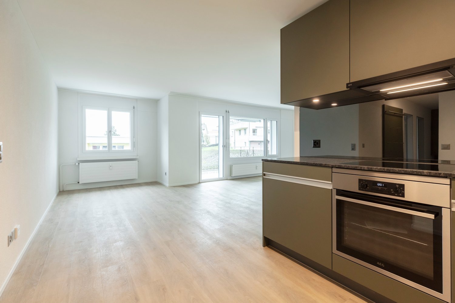Empty kitchen with wooden floor, white walls, stainless steel oven, cabinets, black countertop, modern fixtures.