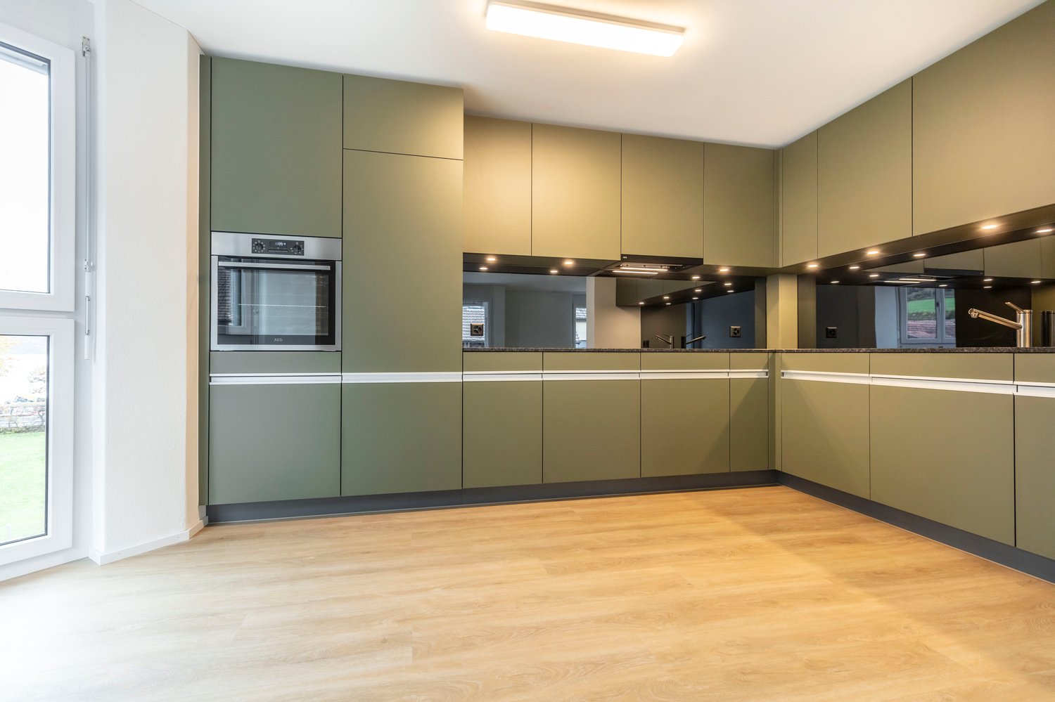 Modern kitchen with sleek green cabinetry, stainless steel appliances, a built-in oven and extractor hood, wooden flooring, large windows, and under-cabinet lighting.