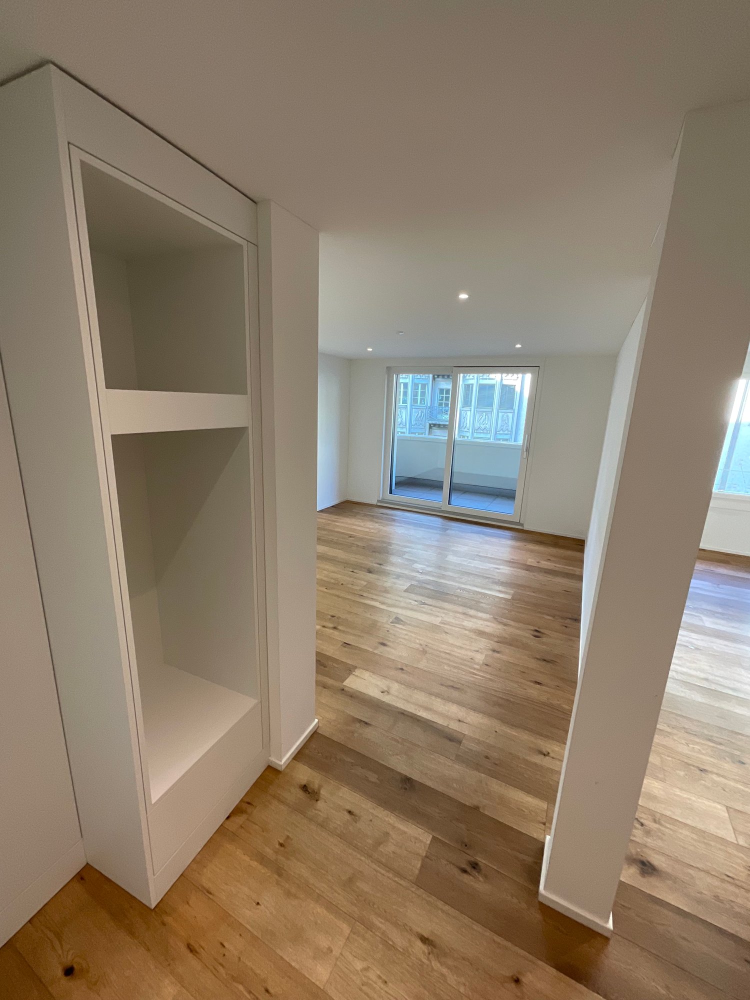 The image shows a spacious room with white walls and a hardwood floor. There is a large window that provides natural light, and the room appears to be empty, with no furniture or decor visible.