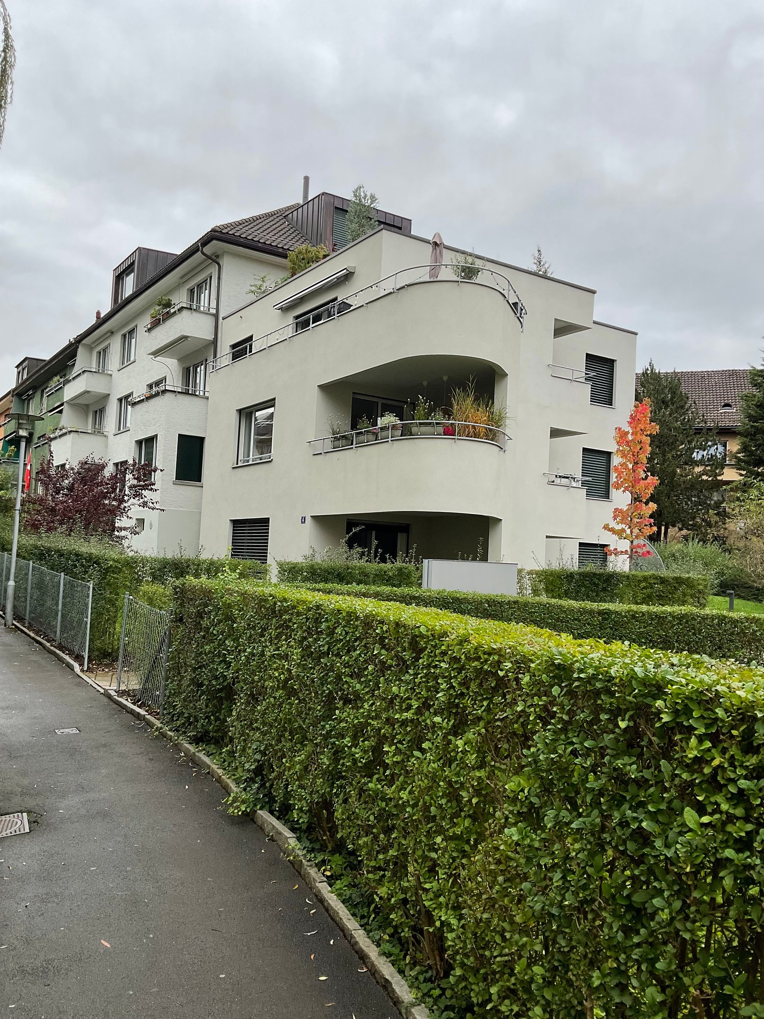Multi-story apartment building with a modern, curved design. The building has several balconies and is surrounded by well-maintained landscaping, including a hedge along the sidewalk.