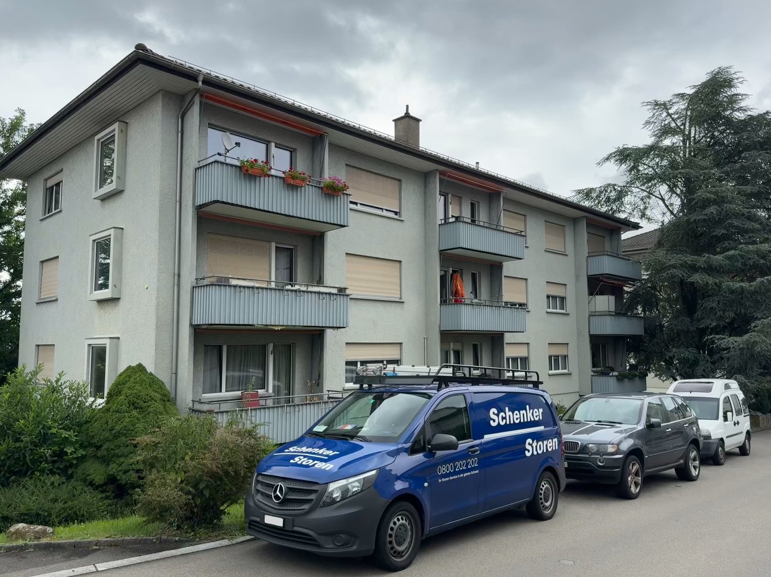 3-story apartment building with gray exterior, balconies, and a Schenker Storen delivery van parked in front