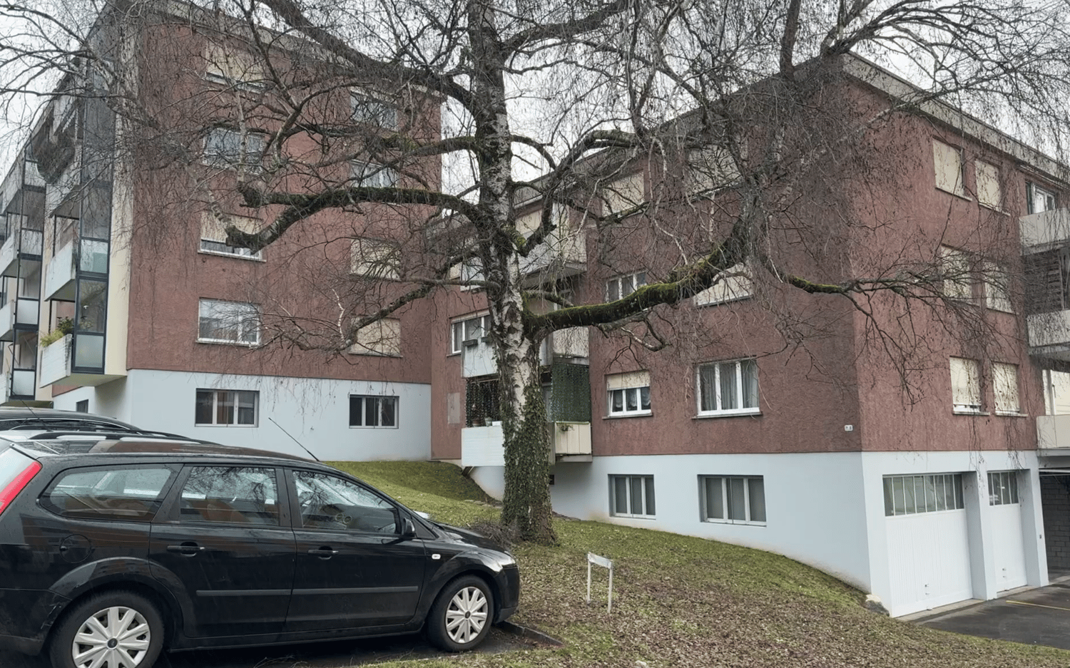 Multi-story building, red brick, white windows, cars parked, large tree in front, grass slope