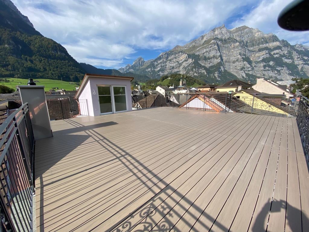 Roof terrace, wooden floor, black railing, panoramic mountain view, a small white building