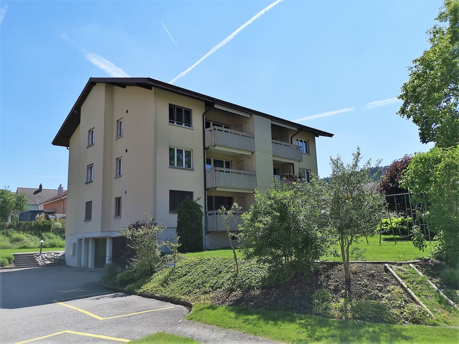 Multi-story building, beige color, multiple balconies, green surrounding area, parking area