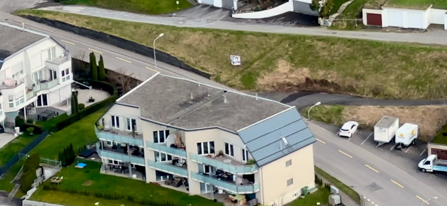 Multi-story apartment building with balconies, surrounded by green space and a road with parked vehicles