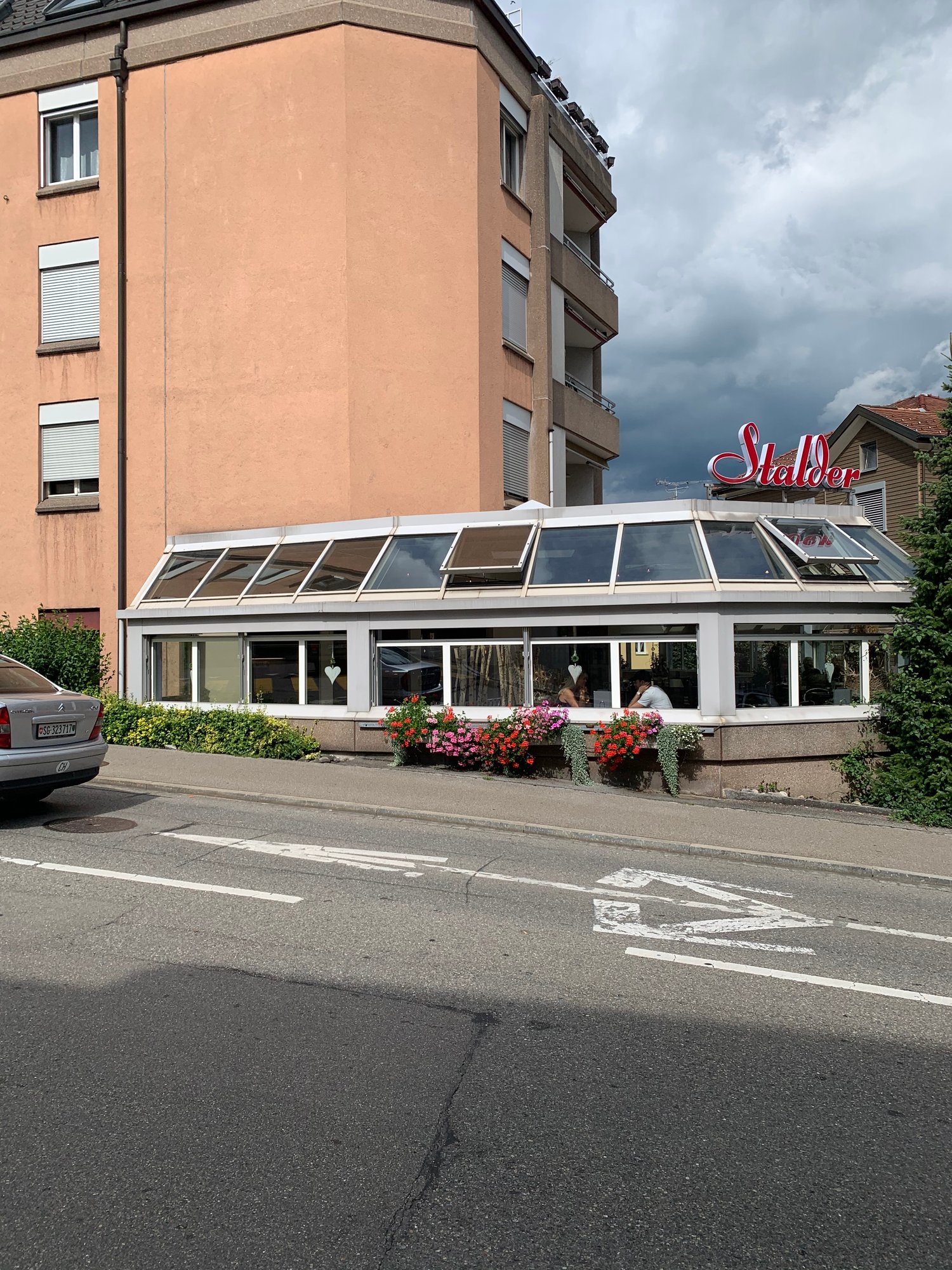 A multi-story building with a large glass-enclosed entryway and a prominent 'Stalber' sign. The building has a peach-colored exterior and appears to be a commercial or residential property located on a street with parked cars.