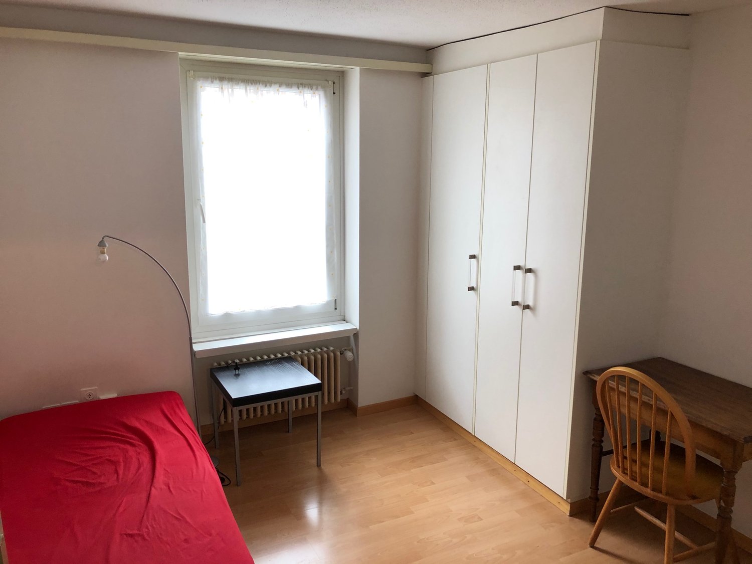 The image shows a bedroom with a large window, a white wardrobe, a red sofa, a small desk, and a wooden chair.