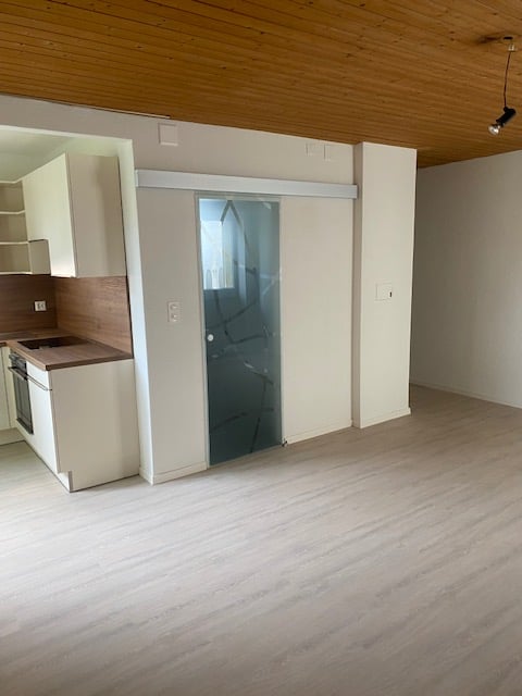 Open-concept studio apartment with hardwood floors, white walls, and a kitchenette with a dishwasher. The room has a sliding glass door leading to another area.