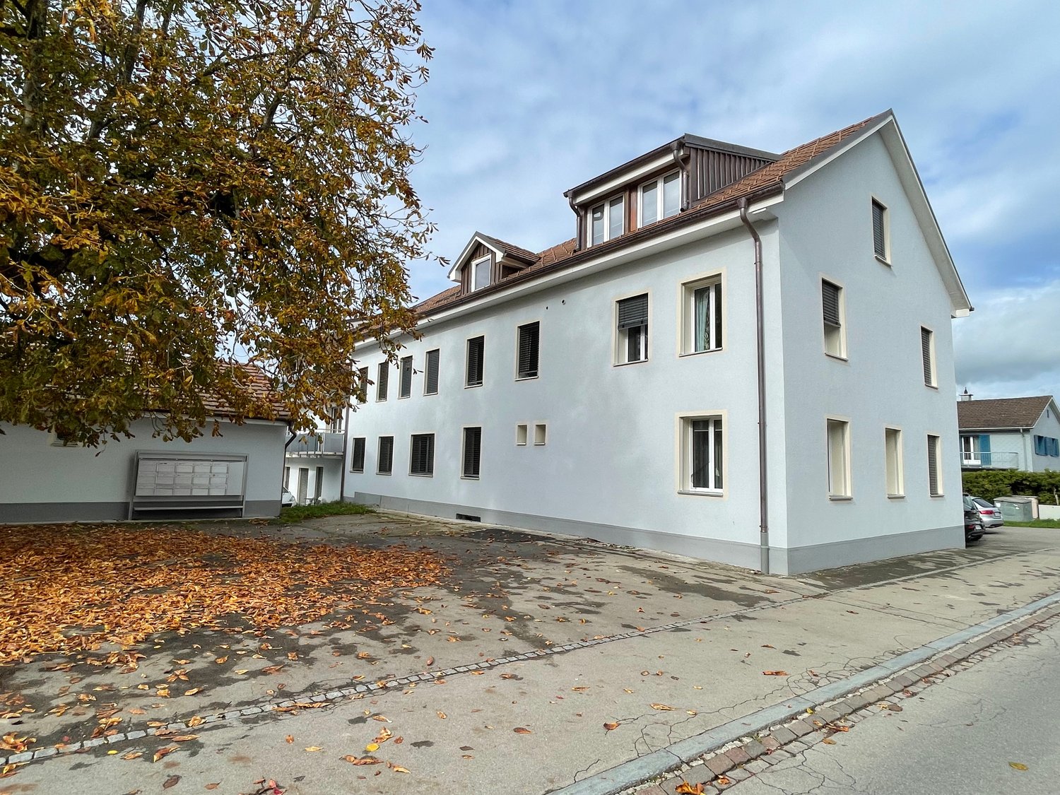 3-story white building, brown roof, multiple windows, large tree in front, parking area, mailbox