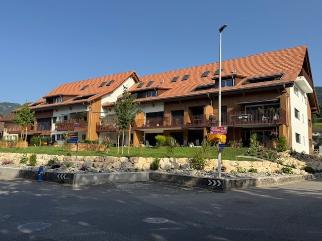 Apartment building, brown roof, beige and wooden exterior, balconies, trees and plants in front