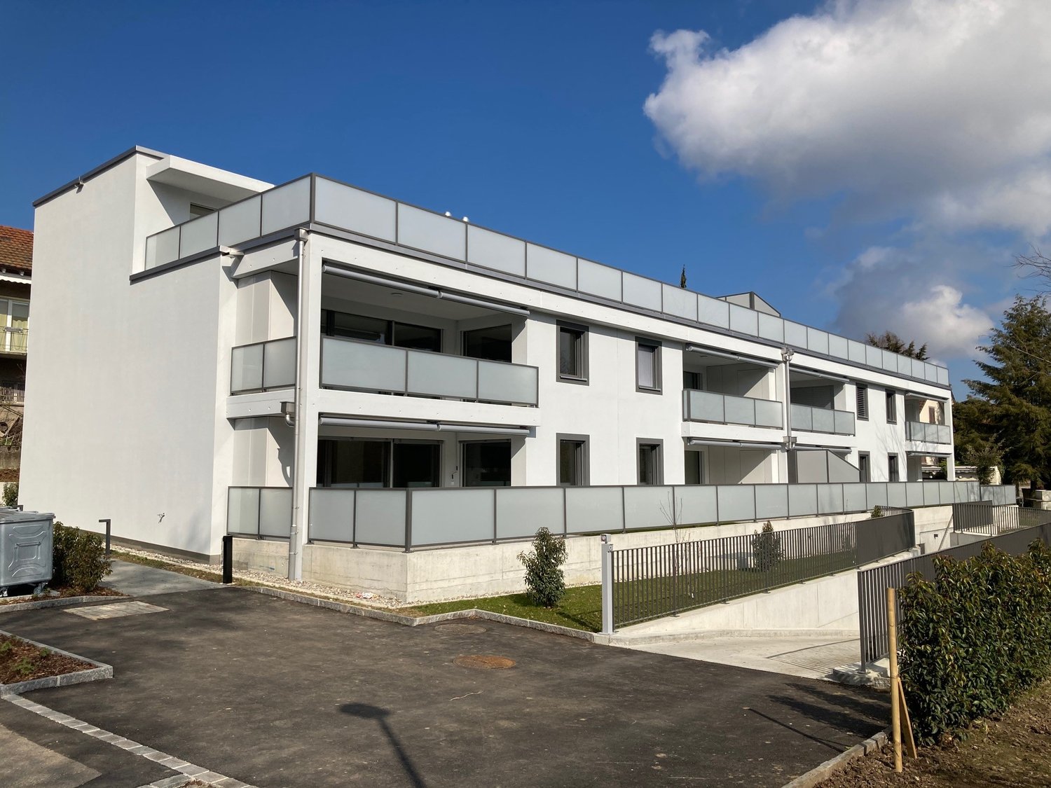 Modern, white apartment building with multiple floors, balconies, and a parking area in front. The ground floor is raised. The entrance is on the right side.