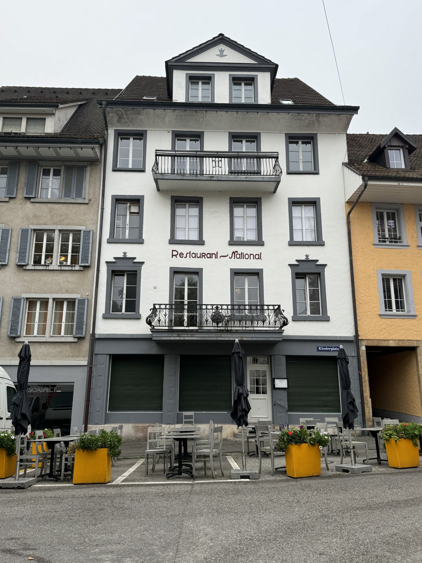 3-story building with a balcony, white exterior, black window frames, and a restaurant called 'Restaurant National' on the ground floor. There are outdoor tables and chairs set up in front of the restaurant.
