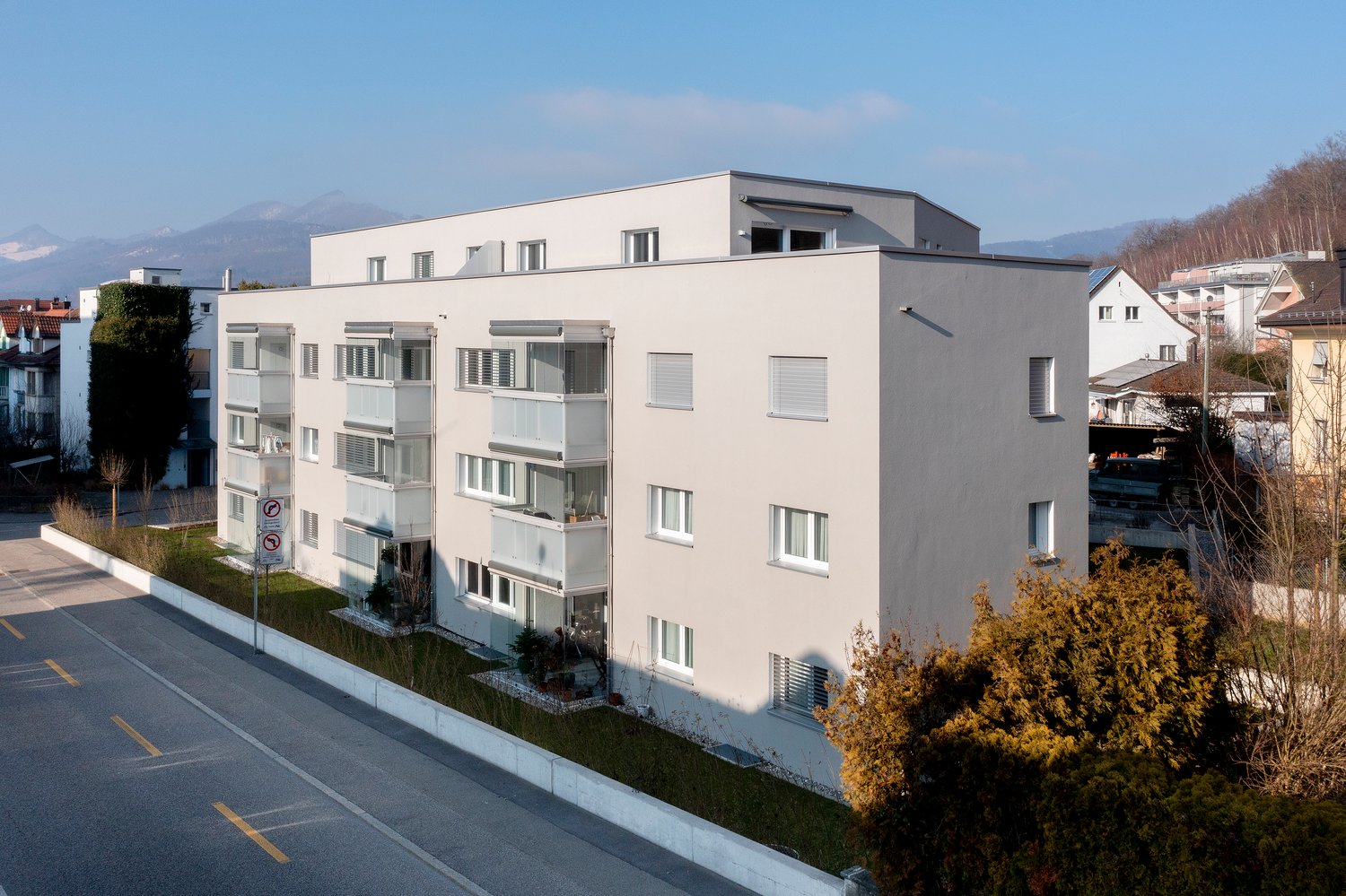 4-story apartment building with white exterior, balconies, and landscaped grounds. The building has a modern, minimalist design with large windows and a flat roof. The surrounding area appears to be residential with some trees and greenery visible.