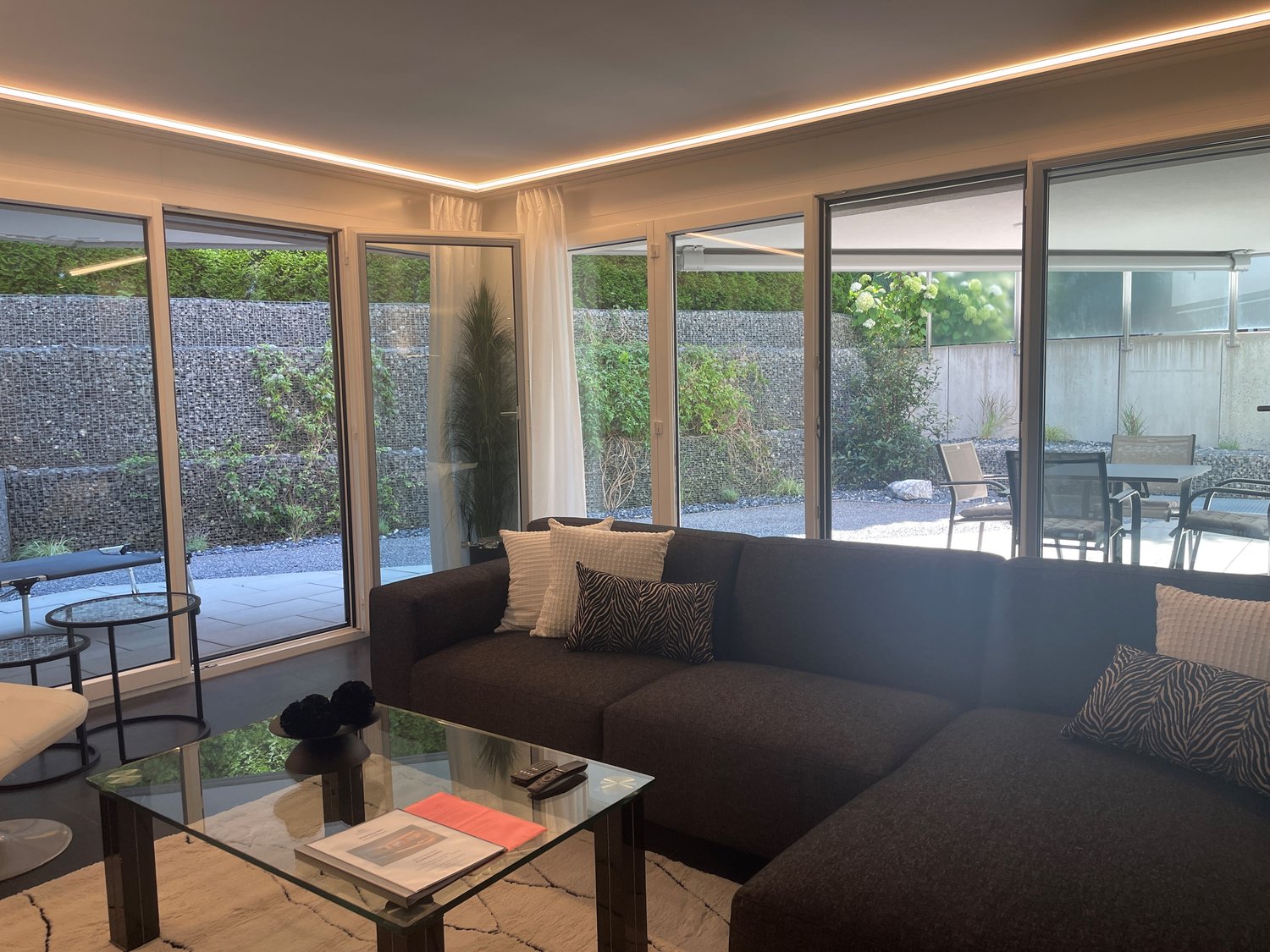 Spacious living room with large windows overlooking a private outdoor area, dark brown sofa, glass coffee table, and decorative pillows
