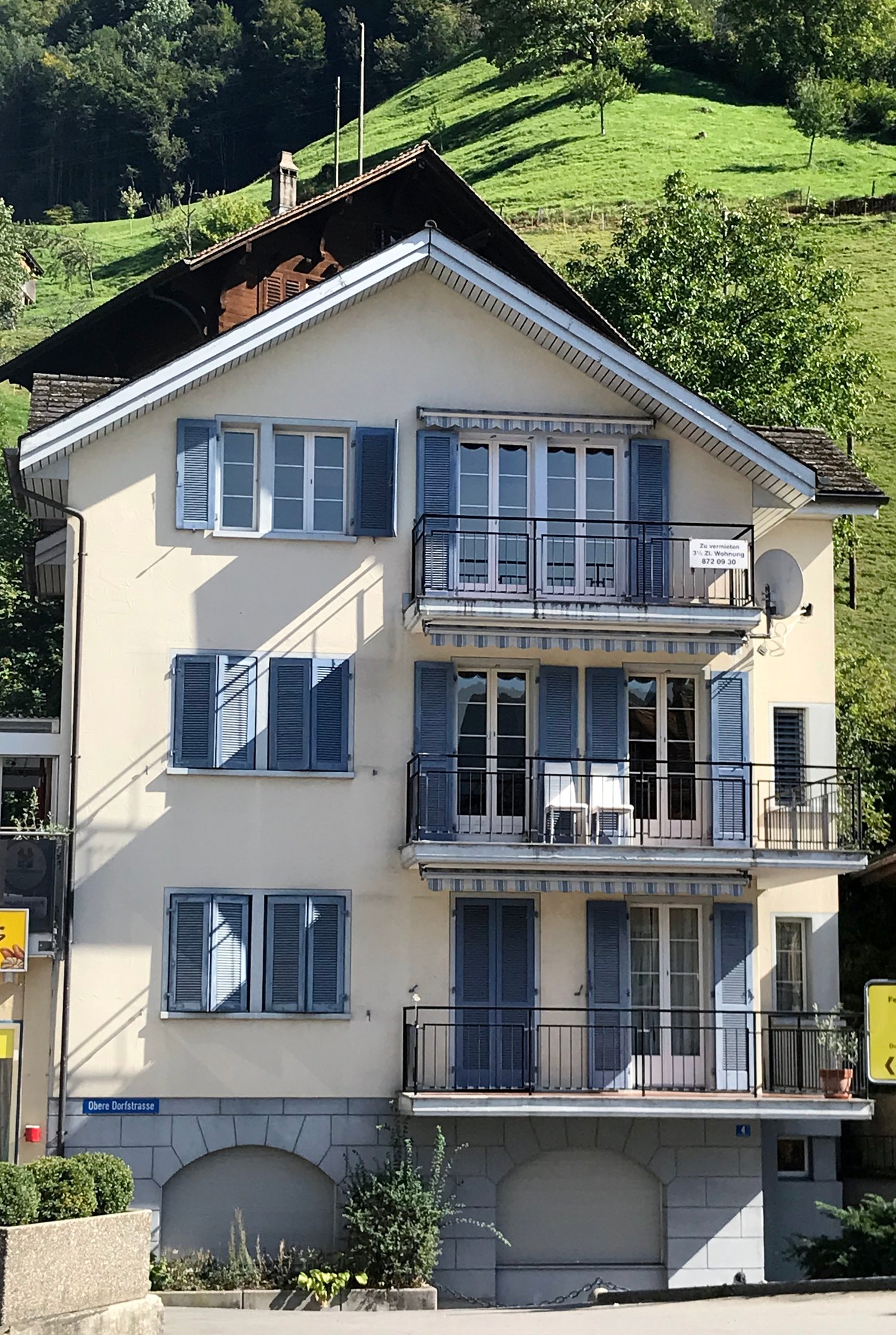 multi-storey house, beige paint, blue shutters, balconies with railings, below another building, garden with plants