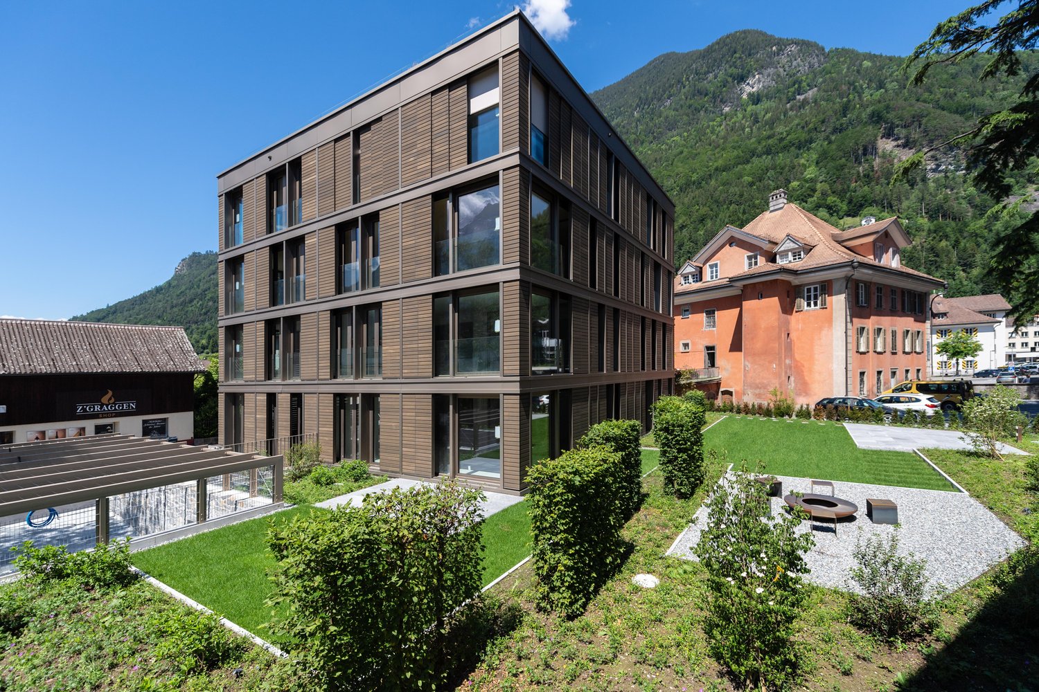 modern building, brown exterior, many windows, greenery, small pathway, other buildings, mountains in the background