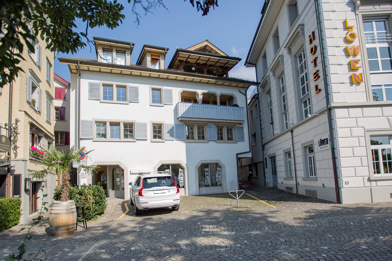 3 story white house with multiple windows, terrace, parking area, flag, plants