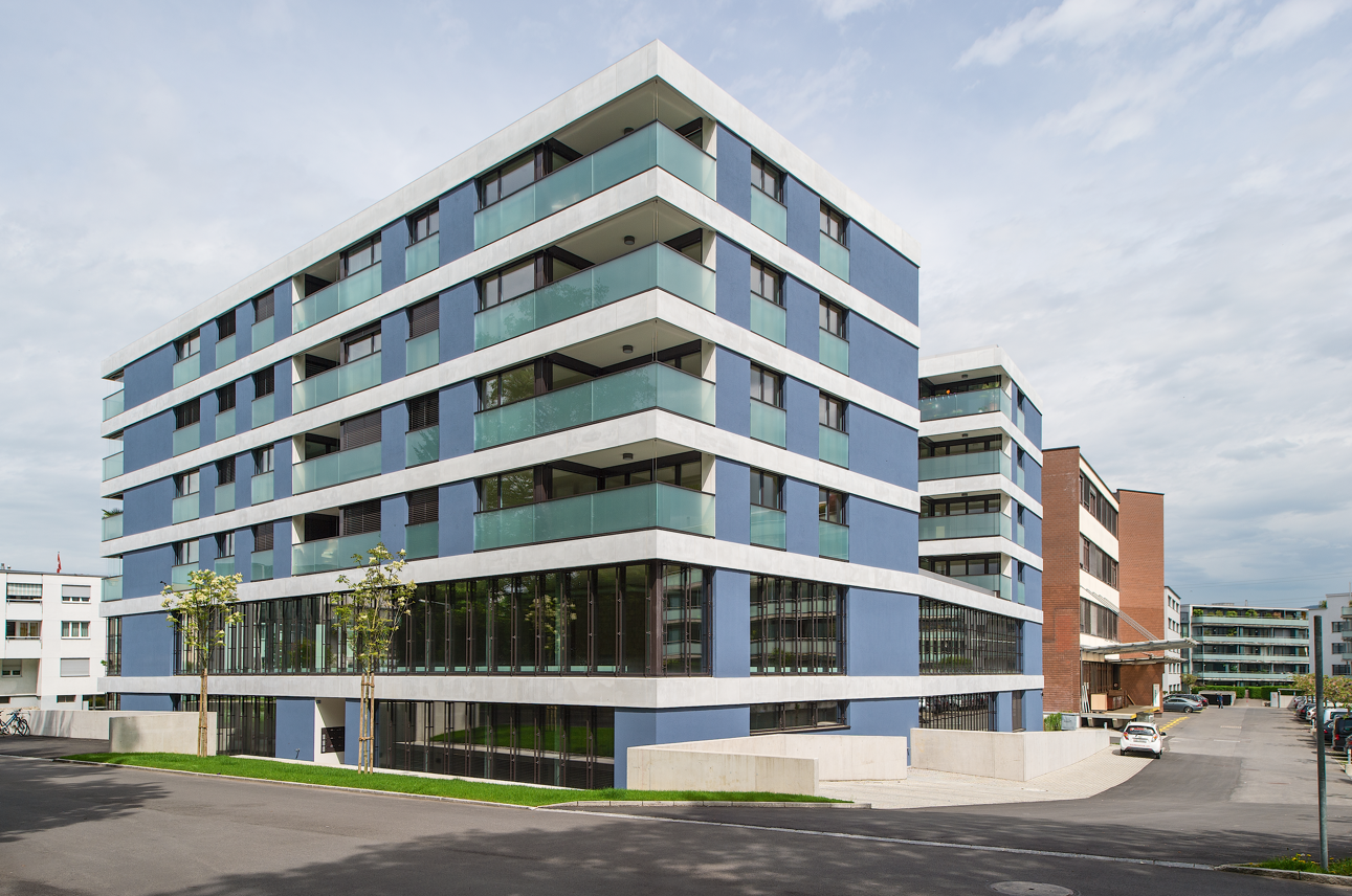 A modern residential building with multiple balconies, blue exterior with white accents, large windows, and is situated on a paved street.