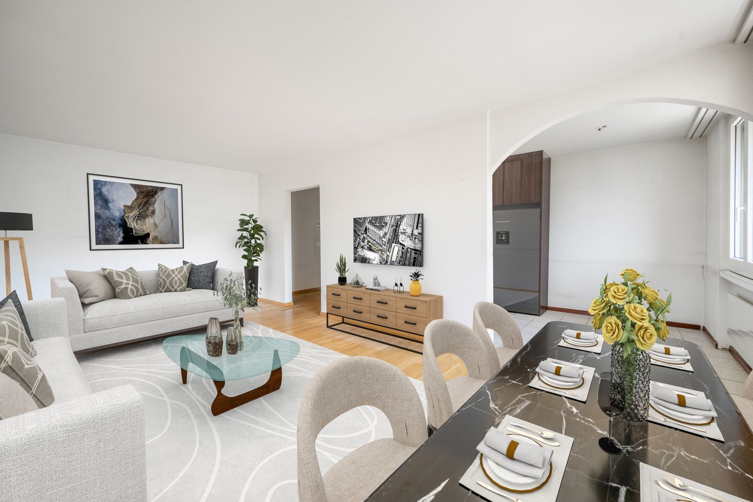 The image shows an open-concept living room and dining room area. The living room features a gray sofa with patterned throw pillows, a glass coffee table, and artwork on the walls. The dining area has a wooden sideboard, a marble-topped dining table with