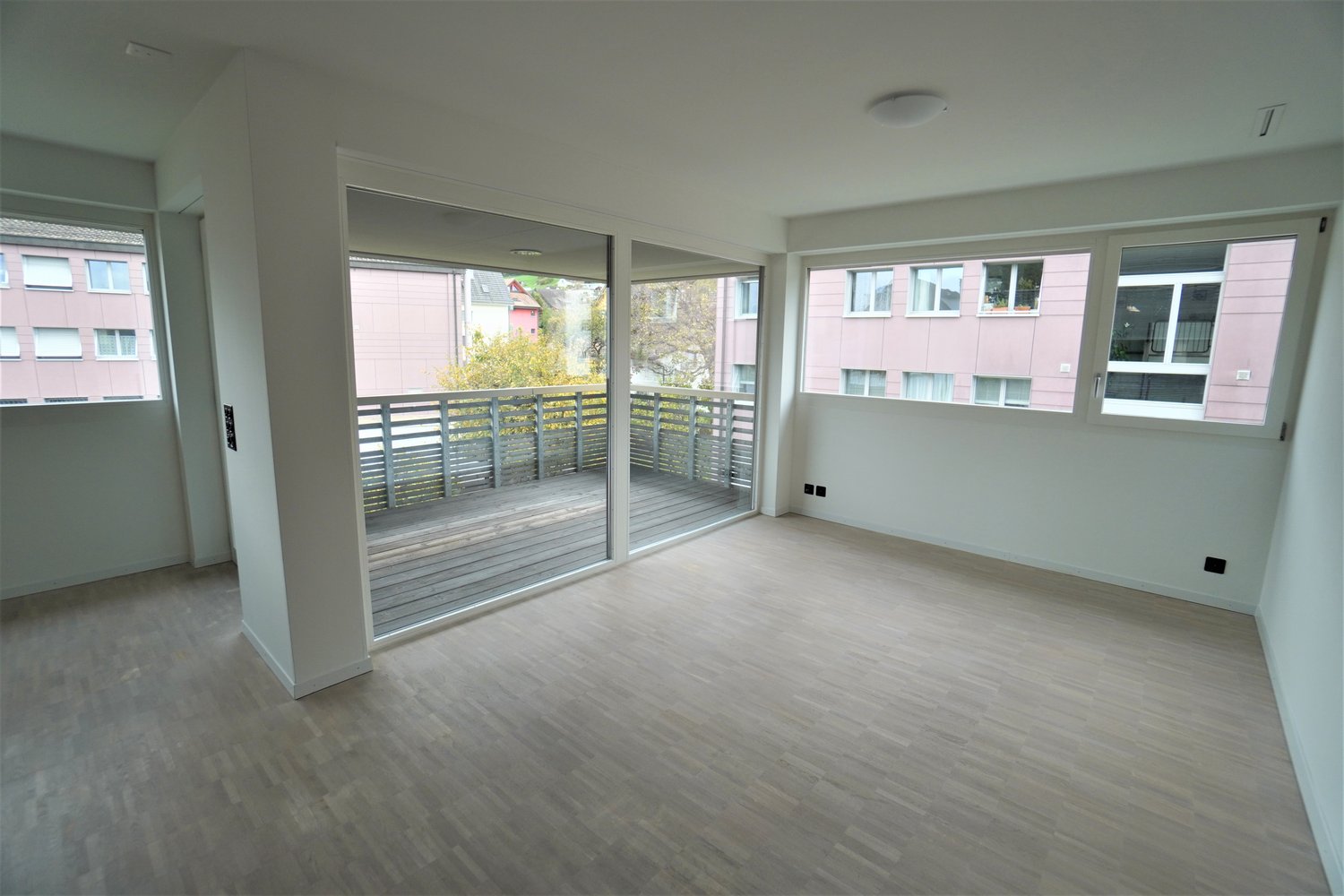 Large windows, sliding glass doors leading to balcony, wooden floors, white walls, power outlets.