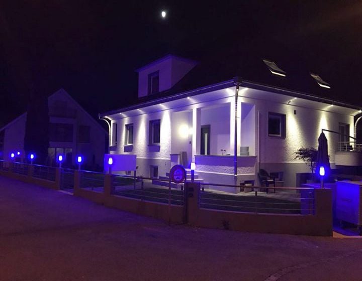 Two-story white house with a balcony, illuminated by purple and white lights at night.